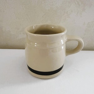 May include: A beige ceramic mug with a rounded barrel shape and a single side handle. The mug features a prominent, solid black horizontal band painted around the lower portion of the exterior body, contrasting with the light cream-colored finish.