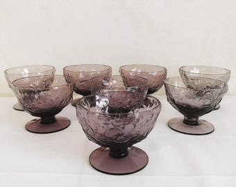 May include: A collection of eight vintage purple glass dessert bowls. Each bowl has a textured surface and sits on a short stem with a round base. The bowls are arranged on a white surface.