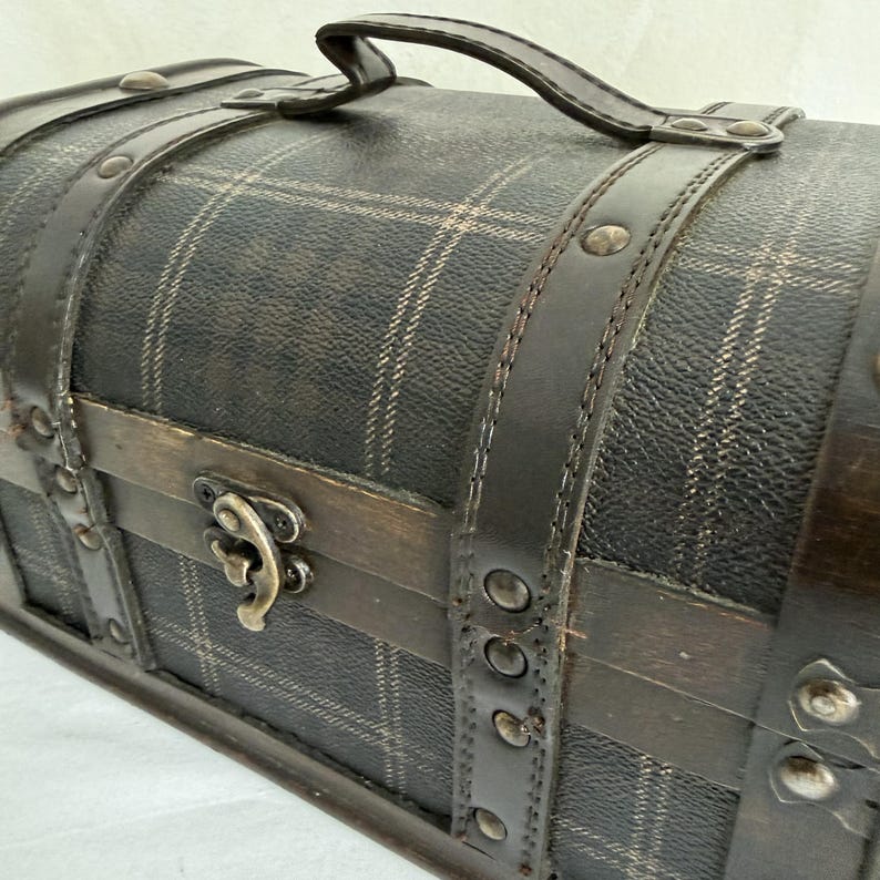 May include: A decorative, vintage-style chest with a curved top. The chest is covered in a dark plaid pattern and features dark brown leather straps and handle. The metal clasp and rivets add to the antique aesthetic.
