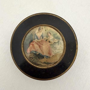 May include: Round, black antique box with a painted scene of two figures in a garden. The central image features a woman in a blue dress and a child, with a pink and peach-colored skirt. The box has a gold trim.