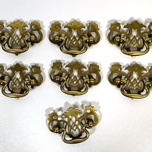 May include: Seven antique brass drawer pulls with a decorative, ornate design. Each pull features a curved handle, two small circular accents, and a central diamond-shaped pattern. The pulls are arranged on a white surface.