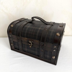 May include: A small, dark brown chest with a plaid pattern, leather straps, and metal studs. It has a curved top, a handle, and a metal clasp. The chest is likely used for storage or decoration.