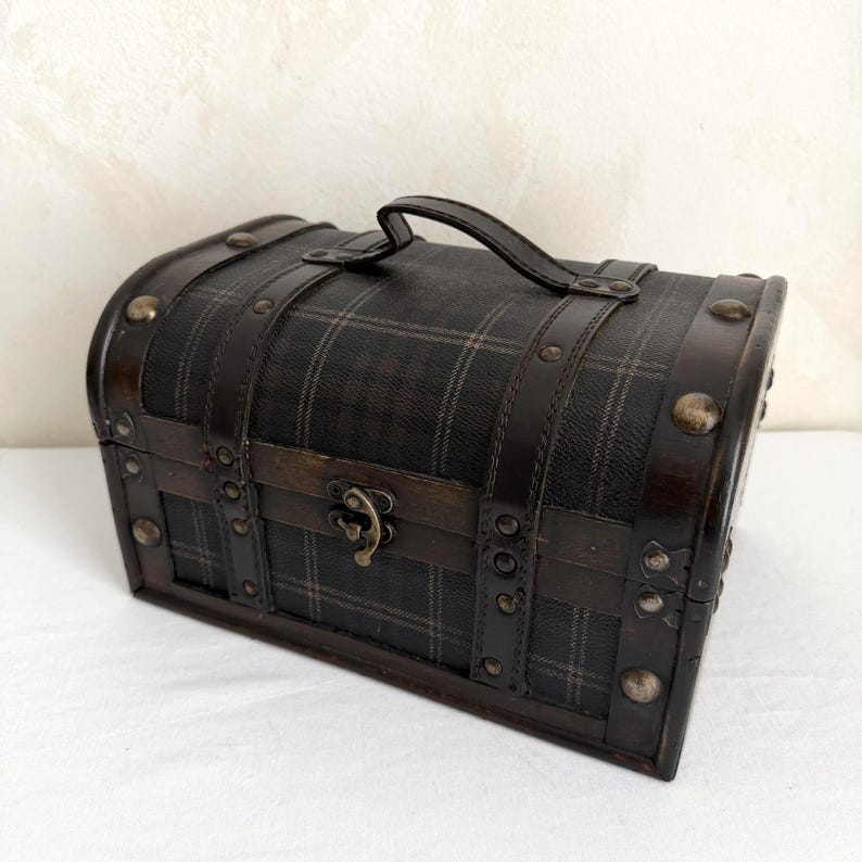 May include: A small, dark brown chest with a plaid pattern, leather straps, and metal studs. It has a curved top, a handle, and a metal clasp. The chest is likely used for storage or decoration.