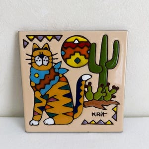May include: A square ceramic tile with a cream background featuring a colorful illustration of a seated cat with a blue bandana, a bright sun, and a green cactus. The design includes geometric patterns in orange, blue, and purple, with the text KRIT.