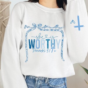 May include: White sweatshirt with the text "she is WORTHY Proverbs 31:7" in blue. The design includes a decorative frame and a cross on the sleeve. The sweatshirt is a casual, comfortable style.