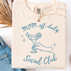 May include: Cream-colored t-shirt with the words "mom off-duty" and "Social Club" in blue script. A blue cocktail graphic is featured below the text. The phrase "EVEN IF IT IS FOR JUST ONE DAY A YEAR!" is printed at the bottom.
