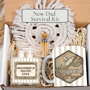 May include: A New Dad Survival Kit in a box, featuring a candle, mug, matches, and a macrame coaster. The mug has a camouflage design with a baby bottle and pacifier graphic, and the name "Marcus" and the year "2026". The candle reads "Deployment in Dad-Ops 2026".