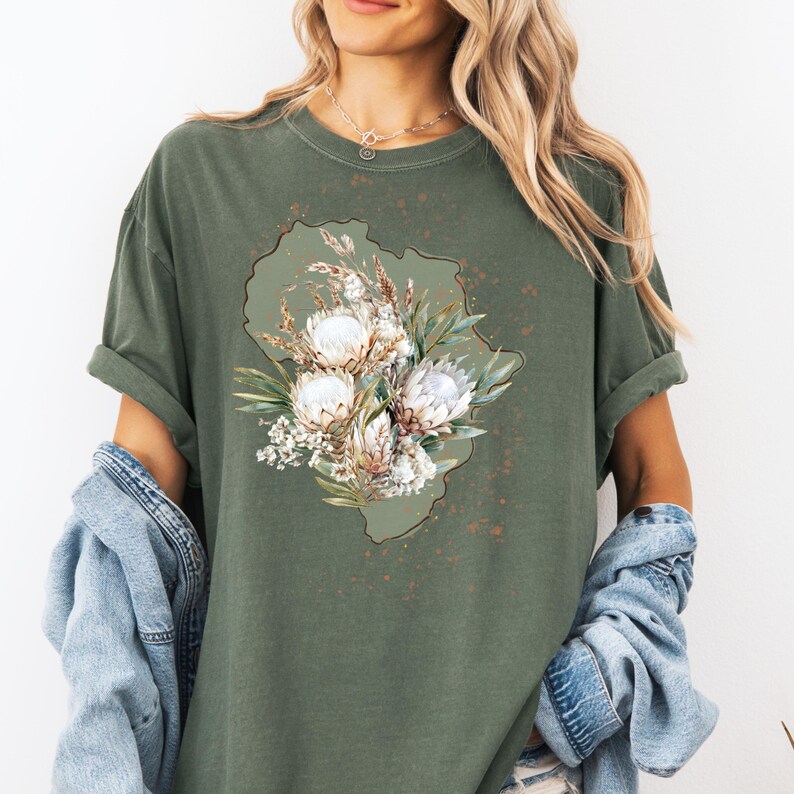 May include: Olive green t-shirt featuring a floral design with white and beige protea flowers, green leaves, and sprigs of wheat. The floral arrangement is set against a green outline. The shirt has rolled-up sleeves and a relaxed fit.