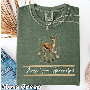 以下が含まれることがあります： モスグリーンのクルーネックTシャツ。植物の中に立つインパラのグラフィックが描かれています。動物の下には、2本の金色のブラシストロークの間に「Always Green. Always Gold」という白い文字がプリントされています。