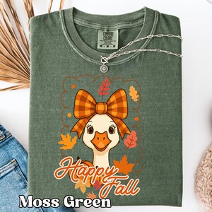 May include: Moss green t-shirt with a graphic of a goose wearing a plaid bow and surrounded by fall leaves. The words "Happy Fall" are written in orange script. A silver necklace with a pendant is draped across the shirt.
