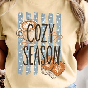 Könnte beinhalten: Ein blassgelbes T-Shirt mit einem Grafikdruck einer Kaffeetasse und einem Paar orangefarbener Fäustlinge. Das Design zeigt blaue vertikale Streifen mit weißen Punkten und den Text COZY SEASON in dunkelbraunen Buchstaben auf der Vorderseite.