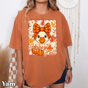 May include: Burnt orange t-shirt featuring a cartoon duck with a plaid bow, surrounded by fall leaves and pumpkins. The text "Happy Fall" is written in a script font. The shirt has short sleeves and a relaxed fit.
