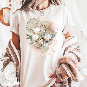 May include: Cream-colored t-shirt featuring a floral design with white and beige flowers, green leaves, and a light green background. The design is centered on the shirt, with a watercolor-style effect. The shirt has rolled-up sleeves.