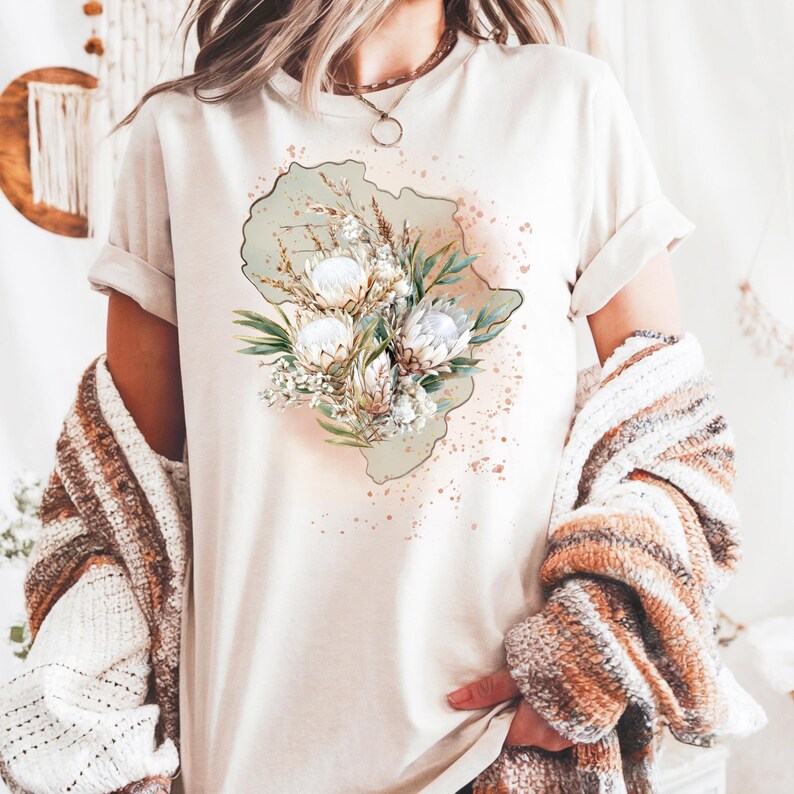 May include: Cream-colored t-shirt featuring a floral design with white and beige flowers, green leaves, and a light green background. The design is centered on the shirt, with a watercolor-style effect. The shirt has rolled-up sleeves.