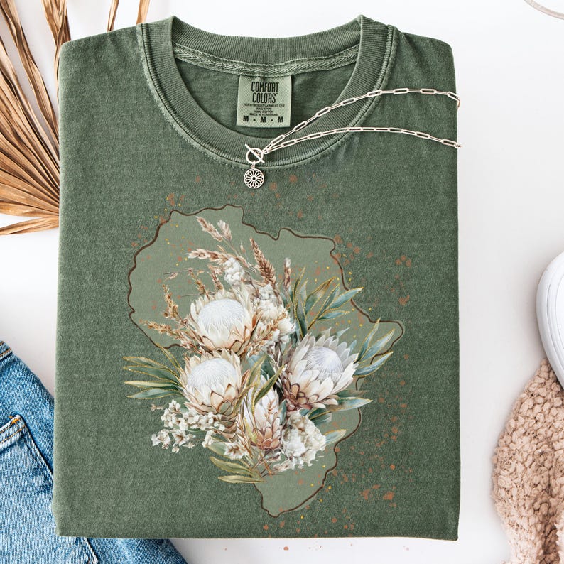 May include: Olive green Comfort Colors t-shirt with a floral design. The design features white protea flowers, wheat, and greenery. A silver necklace with a small pendant rests on the shirt. The shirt is folded and placed on a white surface.