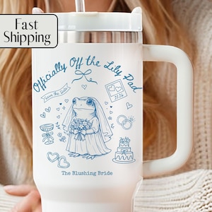 May include: White insulated travel mug with a large handle, featuring a blue illustration of a frog bride with the text "Officially Off the Lily Pad" and "The Blushing Bride". Includes a straw and a lid.