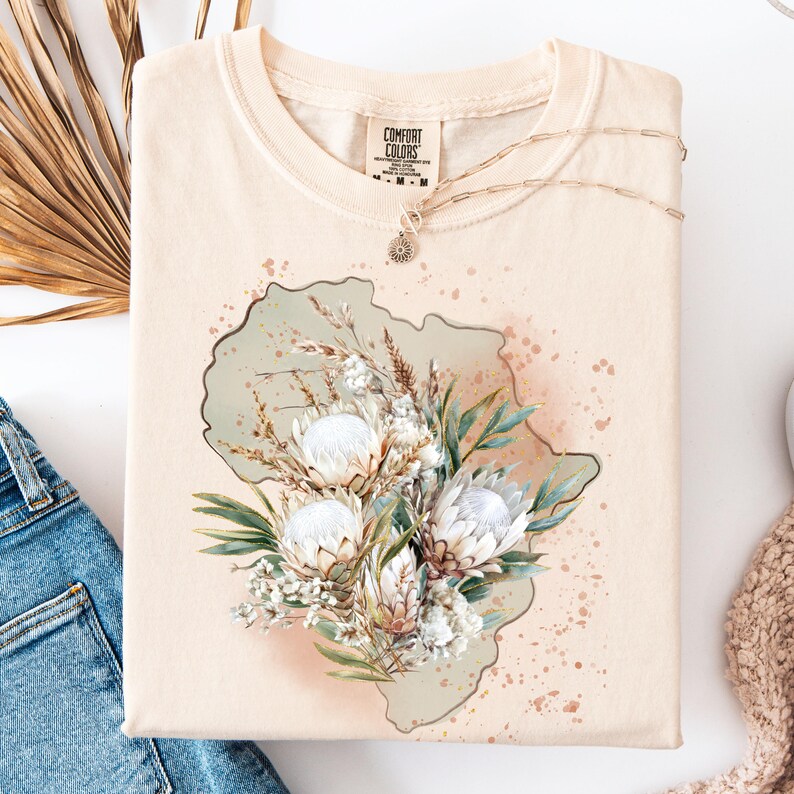 May include: Cream-colored t-shirt with a floral design featuring white protea flowers, green leaves, and dried grasses. The shirt has a crew neck and short sleeves. A delicate gold necklace with a pendant rests on the shirt. The brand name "COMFORT COLORS" is visible.