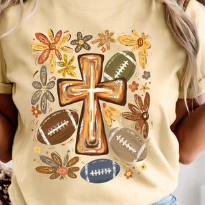 Op de afbeelding: Een lichtgeel t-shirt met een centrale afbeelding van een bruin kruis omringd door diverse gestileerde bloemen in oranje, gele en bruine tinten, afgewisseld met verschillende bruine en blauwe footballs met witte veters.