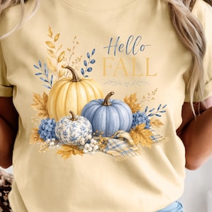 May include: A pale yellow t-shirt featuring a graphic print of three pumpkins in shades of yellow, blue, and white with a blue floral pattern. The design includes autumn leaves, blue flowers, and the text Hello FALL in blue and gold lettering.