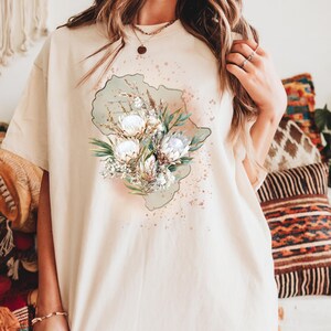 May include: Cream-colored t-shirt featuring a floral design with white and green flowers, and a light green outline. The design includes a watercolor effect with pink and brown accents. The shirt has a relaxed fit.