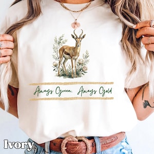 Puede incluir: Camiseta de color marfil con una ilustración en acuarela de una gacela con grandes cuernos, rodeada de vegetación y flores. El texto "Always Green. Always Gold" está impreso en verde debajo. La camiseta es de color neutro.