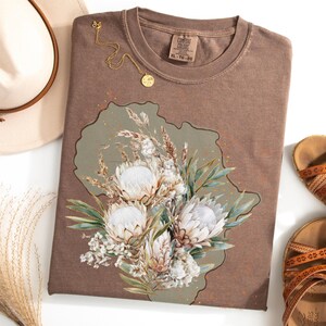 May include: A brown Comfort Colors XL-TG-EG t-shirt with a floral design featuring white protea flowers, green leaves, and dried grasses. A gold necklace and a hat are next to the shirt. Sandals are on the right.