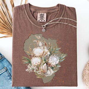 May include: A brown Comfort Colors t-shirt with a floral design. The design features white protea flowers, greenery, and dried grasses on a muted green background. A silver necklace with a pendant rests on the shirt.