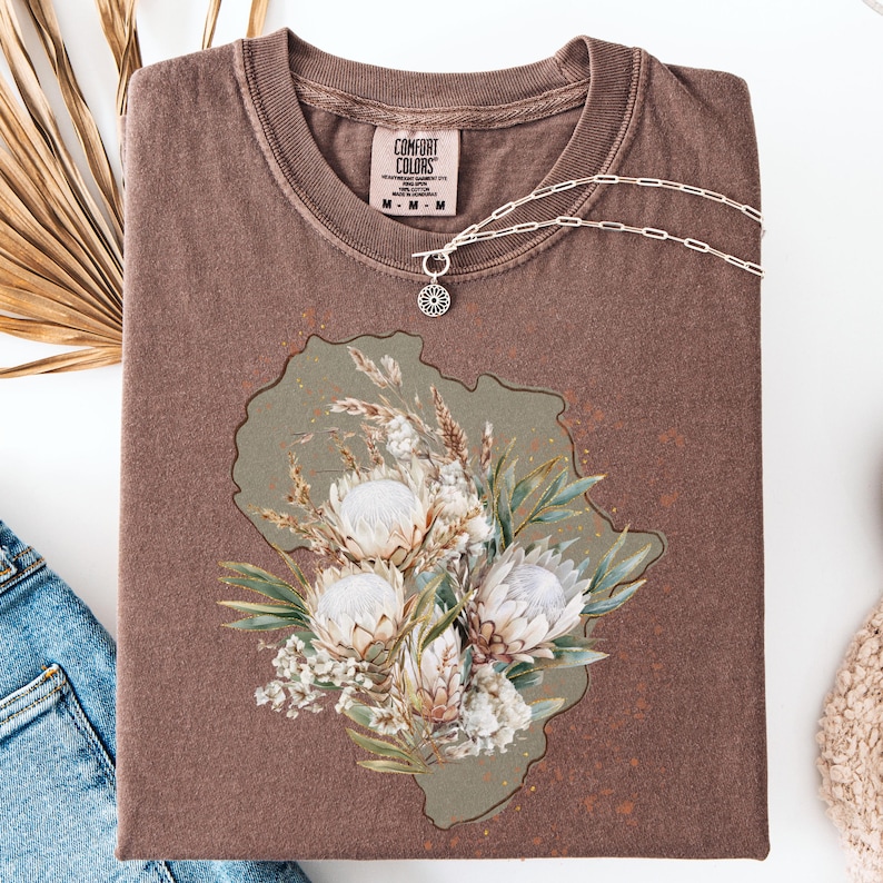 May include: A brown Comfort Colors t-shirt with a floral design. The design features white protea flowers, greenery, and dried grasses on a muted green background. A silver necklace with a pendant rests on the shirt.