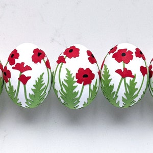 Op de afbeelding: Vijf witte paaseieren versierd met rode klaprozen en groene bladeren. De klaprozen zijn uit papier geknipt en op de eieren aangebracht. De eieren zijn in een rij gerangschikt tegen een witte achtergrond.