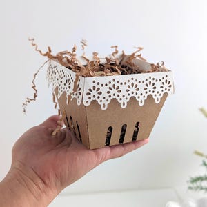 Paper berry basket made from kraft cardstock with a white lace edge detail, held in a hand and filled with crinkle paper as a decorative treat basket.