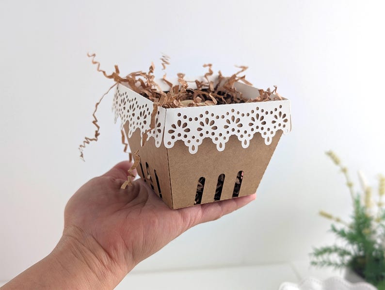 Paper berry basket made from kraft cardstock with a white lace edge detail, held in a hand and filled with crinkle paper as a decorative treat basket.