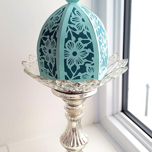 May include: A teal blue paper lantern with a floral design sits on a silver pedestal. The lantern has a domed top and is made of delicate paper.