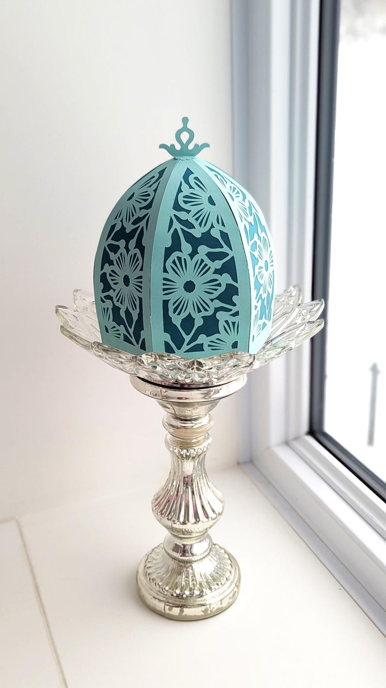 May include: A teal blue paper lantern with a floral design sits on a silver pedestal. The lantern has a domed top and is made of delicate paper.