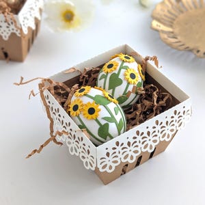 Lace-edge paper berry basket made from kraft cardstock, filled with shredded paper and two sunflower-decorated Easter eggs, styled on a white background for a Lace Easter Basket SVG treat holder template for Cricut and cutting machines.