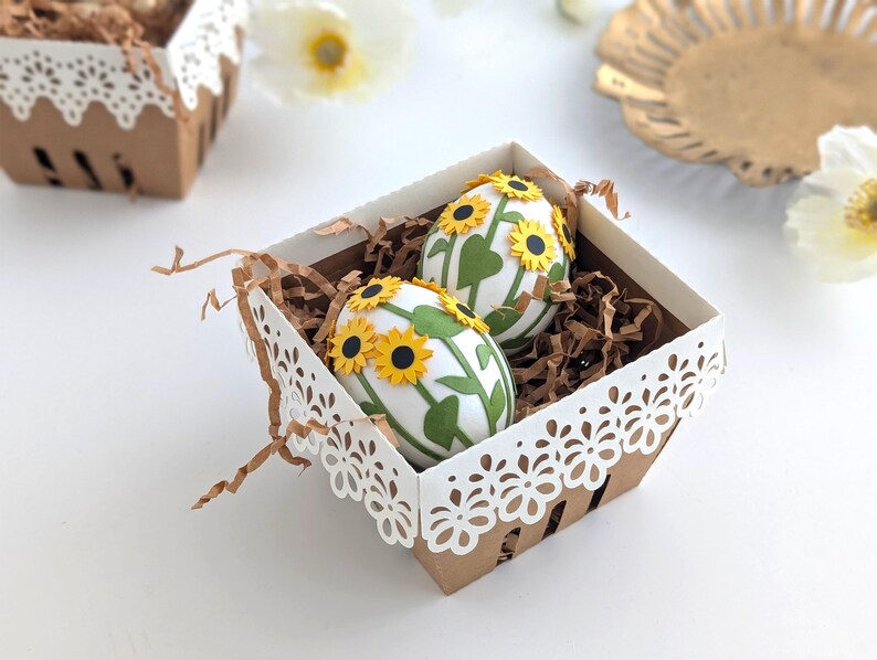 Lace-edge paper berry basket made from kraft cardstock, filled with shredded paper and two sunflower-decorated Easter eggs, styled on a white background for a Lace Easter Basket SVG treat holder template for Cricut and cutting machines.