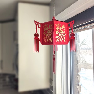 Red paper Chinese lantern with floral cutouts and tassels hanging by a window, created from an SVG file for Lunar New Year home décor.