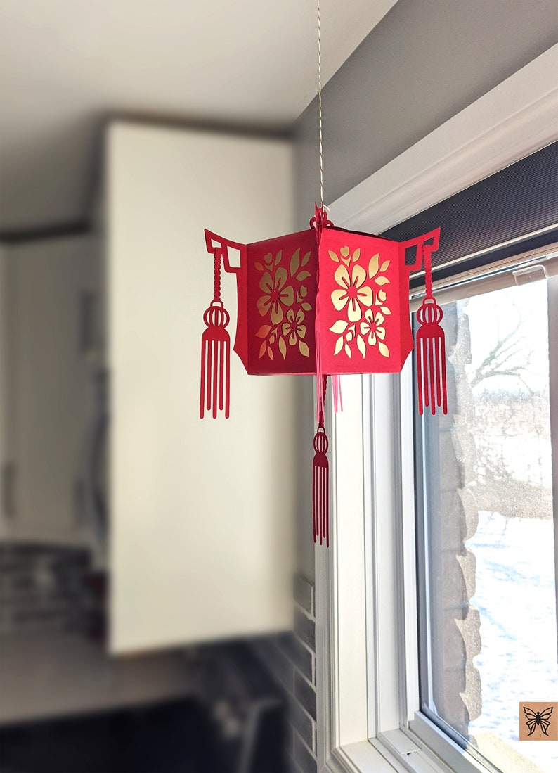 Red paper Chinese lantern with floral cutouts and tassels hanging by a window, created from an SVG file for Lunar New Year home décor.