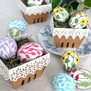 Decorative paper Easter egg wraps in pastel leaf, sunflower, and poppy designs displayed in white lace berry baskets on a spring table setting.