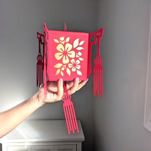 Hand holding a red paper Chinese lantern with gold floral cutouts and tassels, made from a 3D SVG file for Cricut and Lunar New Year décor.