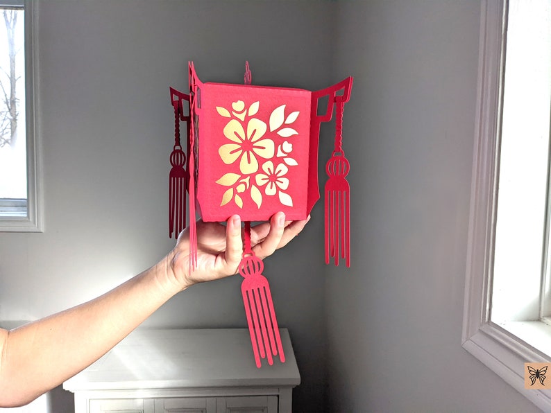 Hand holding a red paper Chinese lantern with gold floral cutouts and tassels, made from a 3D SVG file for Cricut and Lunar New Year décor.