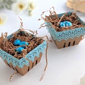 Two lace-edge paper berry baskets in blue and kraft cardstock, filled with shredded paper and blue chocolate eggs, styled on a white background with soft spring flowers for a Lace Easter Basket SVG treat holder template for Cricut and cutting machines.