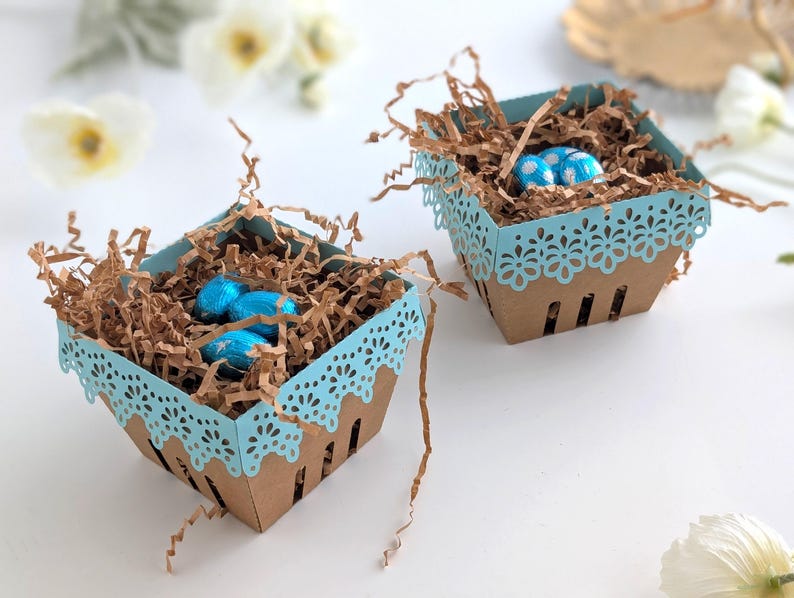 Two lace-edge paper berry baskets in blue and kraft cardstock, filled with shredded paper and blue chocolate eggs, styled on a white background with soft spring flowers for a Lace Easter Basket SVG treat holder template for Cricut and cutting machines.