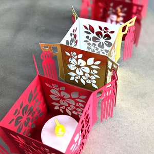 Row of red and gold paper Chinese tea light holders with floral cutouts and tassels, each crafted from SVG templates and lit with small LED candles.
