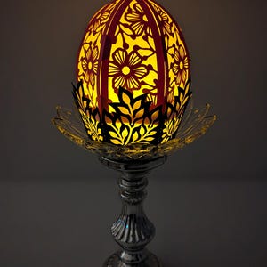 May include: A decorative paper egg with a floral design, lit from within, sits on a silver stand. The egg is pink and white with a black outline. The stand is a classic design with a round base and a tall, slender stem.