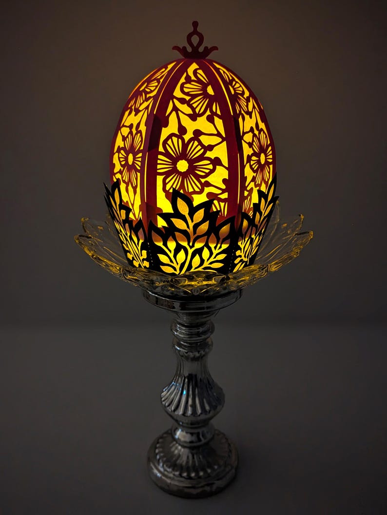 May include: A decorative paper egg with a floral design, lit from within, sits on a silver stand. The egg is pink and white with a black outline. The stand is a classic design with a round base and a tall, slender stem.