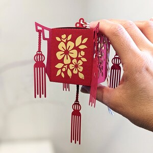 Hand holding a small red paper Chinese lantern with floral cutouts and tassels, crafted from an SVG template for Cricut and Lunar New Year decorations.