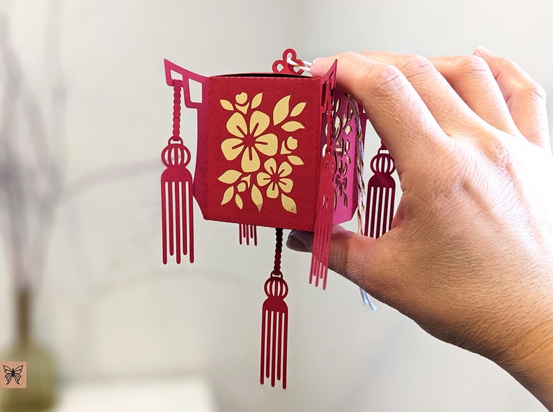 Hand holding a small red paper Chinese lantern with floral cutouts and tassels, crafted from an SVG template for Cricut and Lunar New Year decorations.