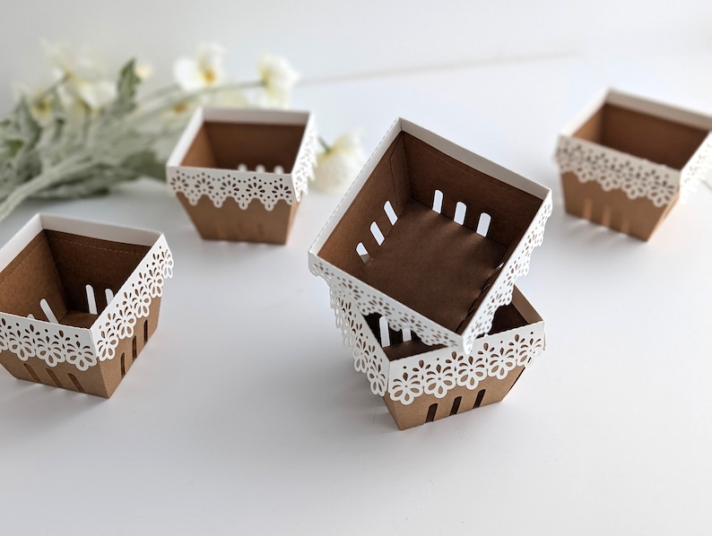Five lace-edge paper berry baskets made from kraft cardstock with white floral lace rims, arranged on a white background with soft spring flowers, shown as a Lace Berry Basket SVG treat holder and favor box template for Cricut and cutting machines.