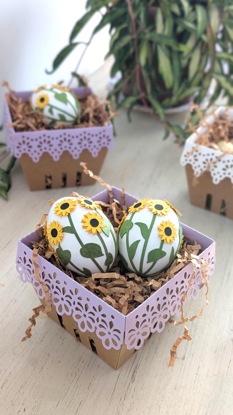 Lavender lace-edge paper berry basket in kraft cardstock filled with shredded paper and sunflower-decorated Easter eggs, styled on a light wood surface with additional baskets in the background for a Lace Easter Basket SVG treat holder template for Cricut and cutting machines.