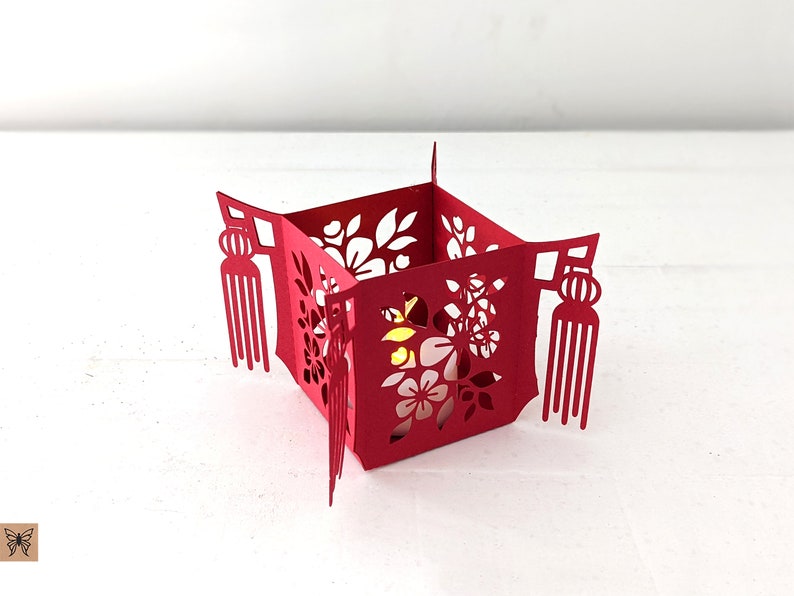 Red paper Chinese tea light holder with floral cutouts and tassel accents, made from a 3D SVG template and illuminated with a small LED light.